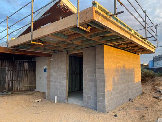 A partially constructed Changing Places facility with concrete block walls and timber scaffolding overhead. The building is on a sandy site, and the interior framing is visible through the open doorway.